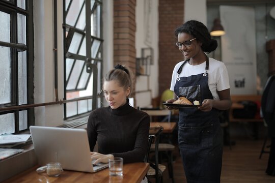 Waitress serving croissant to woman working on laptop