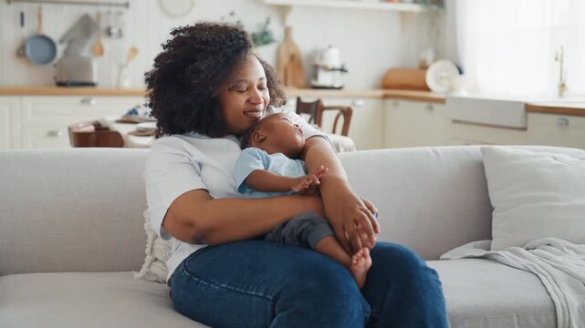 Happy african woman holding her baby in hands and resting in sofa in living room . Portrait of mother and child in cozy home interior, positive emotions, feel happiness, joy and tenderness, family