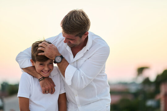 Father Playfully Hugging Son On Balcony Overlooking Sea At Sunset Family Bonding Moment