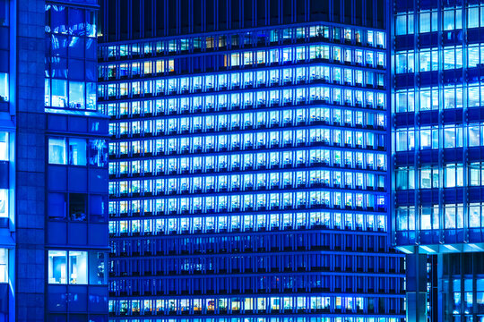 High-density facade of modern illuminated office buildings in Tokyo at night with geometric blue glass windows.