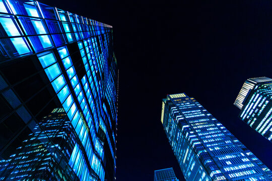 Dramatic bottom-up looking perspective of futuristic corporate skyscrapers in Tokyo at night with glowing blue windows.