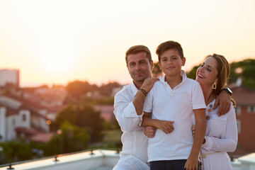 Obraz premium Happy Family On Rooftop Balcony At Sunset Over Coastal Town With Parents Embracing Son