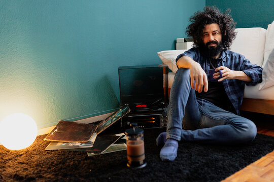 Bearded man with tangled hair holding coffee cup while sitting by turntable in living room