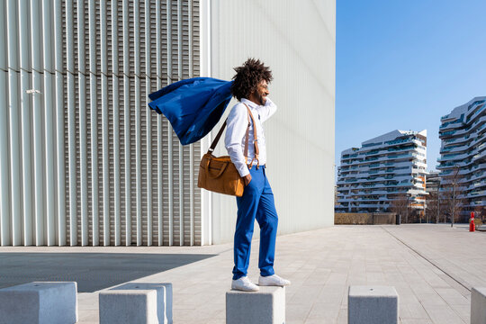 Smiling businessman holding blazer on shoulder standing with shoulder bag on concrete block