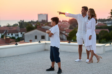 Obraz premium Family On Rooftop Terrace At Sunset With Sea View, Parents Pointing While Son Watches