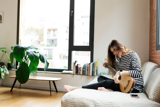 Happy young woman sitting on couch at home playing guitar