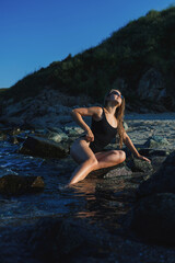 Fototapeta premium Person in black swimsuit poses on rocks by water during daylight near a beach on a sunny day with clear blue sky and greenery in the background