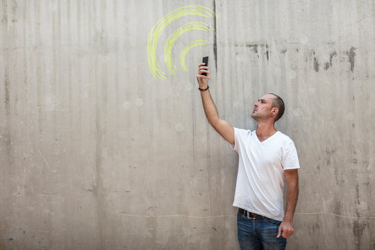Man holding up phone looking for signal with wifi sign drawn in yellow chalk on concrete wall