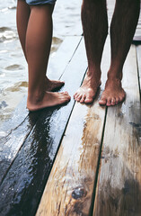 Fototapeta premium Two individuals, one with their foot higher and the other lower, touch the damp boards near the shoreline as they relax on the wooden walkway. Summer day enjoyment by the dock.