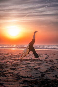 Woman lifting one leg high in the three legged downward dog yoga pose on the sand as the sun sets over the sea a dynamic posture of strength balance and outdoor energy.