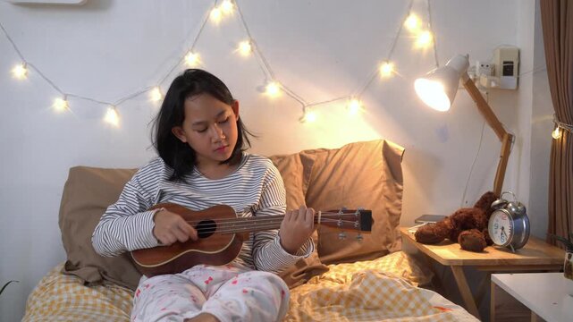 Young girl sits comfortably on her bed practicing the ukulele after her online classes in bedroom illuminated by warm fairy lights