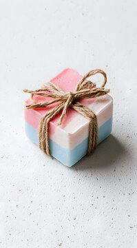 A handmade soap bar featuring pastel pink and blue layers, tied with natural twine. Captured in a simple, minimalist product shot against a clean white speckled background. Luxurious for spas and bath