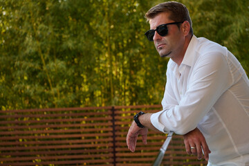 Obraz premium Relaxed Man On Home Balcony Wearing Sunglasses Leaning On Railing Enjoying Summer Rooftop View