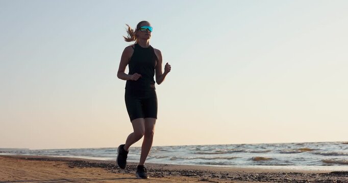 Sporty woman jogging on Baltic sea beach at sunset in black sportswear. Healthy active lifestyle and fitness. Cardio workout