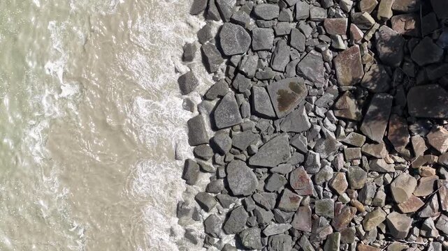 Waves crash against jagged rocks below clear sky. Water swirls with motion, hitting stone surface