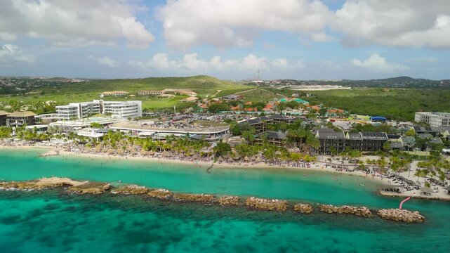 Aerial parallax over Mambo Beach Curacao reveals packed sandy shore turquoise water rocky breakwater palm trees and large resort complex against green hills