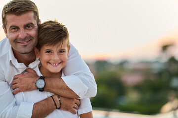 Obraz premium Father Hugging Smiling Son On Balcony At Sunset Overlooking Sea View From Rooftop