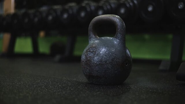 A kettlebell rests on the floor of a gym surrounded by various weights. The focus is on strength training, suitable for different fitness levels