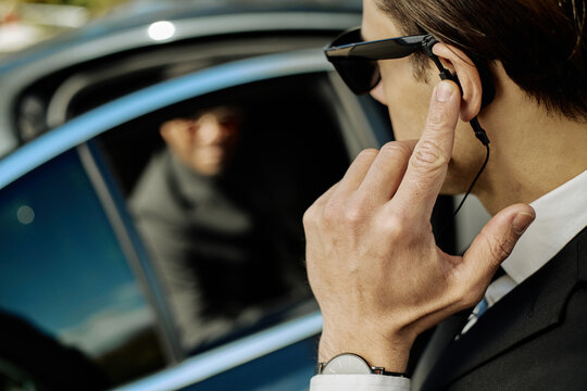 Close up of young adult Caucasian man adjusting earpiece while guarding client near car, conveying executive protection. Ideal for security service, VIP transport, risk management marketing