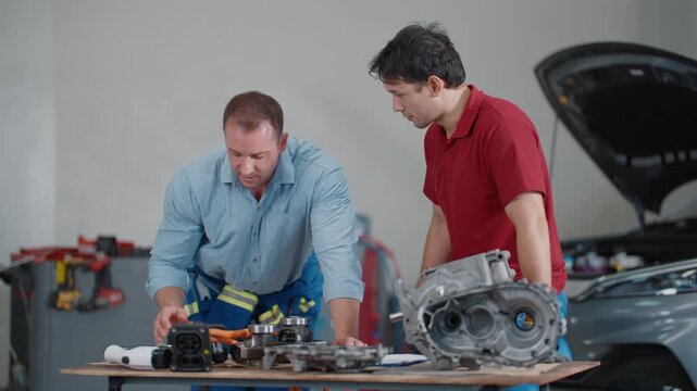 Two mechanics examine engine parts at a repair shop. One gestures while speaking to his colleague. They work beside a lifted car, tools scattered nearby. Teamwork and technical focus