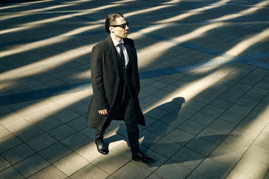 High angle view of Caucasian young adult businessman walking across urban plaza, wearing sunglasses and commuting with confidence. Ideal for leadership, career growth, corporate branding