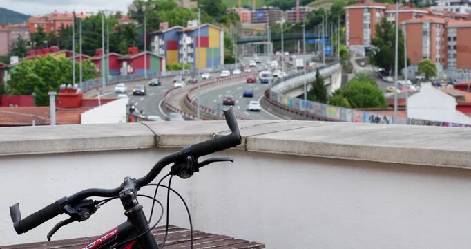 Bicycle and Urban Highway View: A bicycle parked on a rooftop terrace overlooks a busy multi-lane urban highway with moving traffic and apartment buildings in the background.