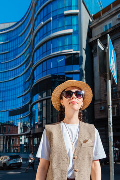 Portrait of a young woman in a straw hat standing in front of a blue glass skyscraper. Vertical