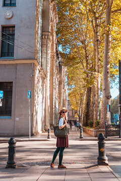Young woman standing on a street lined with tall autumn trees and a stone building. Vertical