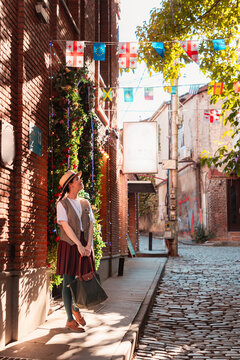Woman standing by a red brick wall under Georgian flags and fairy lights. Vertical