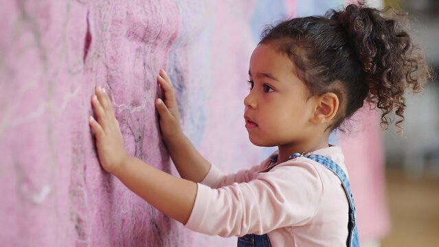 Profile view of young girl touching soft pink fiber art installation on wall showing interest in tactile sensations and creative textures while visiting gallery or educational center.