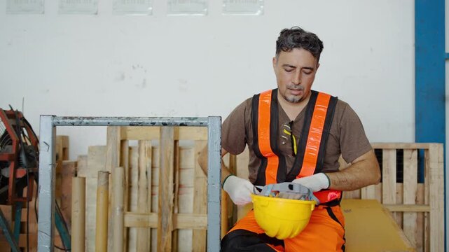 Exhausted middle aged male construction worker taking a well-deserved break at an industrial site. He removes his yellow hard hat, wipes sweat from his forehead, and rests after heavy physical labor.