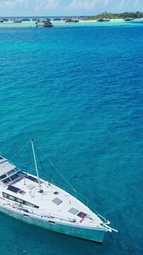 Close aerial sailboat on turquoise lagoon single sailing vessel gliding over clear shallow coral waters captain tending lines sunlight sparkling on hull calm breeze ideal, Maldives, vertical video