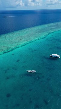 Aerial view turquoise reef with two boats anchored over shallow coral deepblue horizon sunlit water distant speedboat leaving faint wake tranquil atmosphere ideal for snorkeling, Maldives
