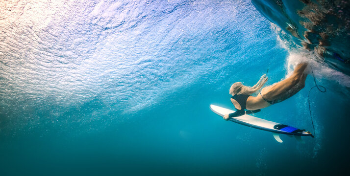 Underwater view of female surfer diving deep with board in crystal clear blue ocean waters