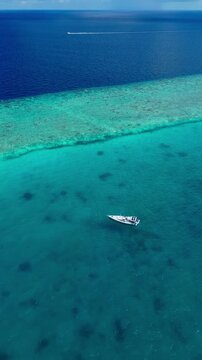 Close aerial sailboat on turquoise lagoon single sailing vessel gliding over clear shallow coral waters captain tending lines sunlight sparkling on hull calm breeze ideal, Maldives, vertical video