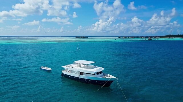 Aerial view yacht anchored turquoise lagoon overlooking coral reef and tiny island panoramic drone shot captures luxury vessel with tender moored nearby, crystal clear shallow,Maldives
