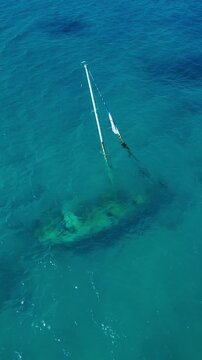 Aerial view sunken yacht in turquoise water, clear visibility revealing leaning mast and halfburied hull, coral patches on seabed, slow current and cinematic drone sweep, survey, vertical video