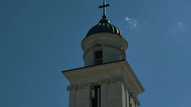Low-angle view of classical bell tower with domed cupola, cross, carved capitals, dentil cornice, pilasters, and rounded arch against clear blue sky with strong daylight and clean composition