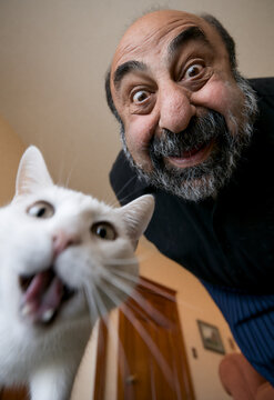 Humorous low-angle photo of stunned bearded man and white cat reacting with surprise as they lean toward the camera to inspect something, close-up blur enhancing lively and spontaneous indoor moment