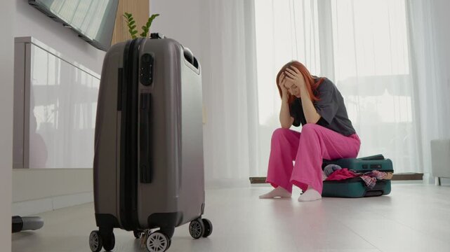 A woman sits on a suitcase with clothes around her. She holds her head in her hands, feeling stressed as she thinks about preparing for her upcoming trip.