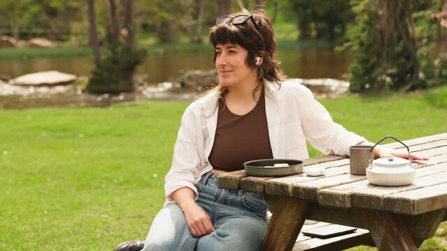 Woman sits at a wooden picnic table. Green grass and a lake surround her. She wears a white cardigan and jeans. She holds a teapot, looking toward the water. Her expression is calm and serene