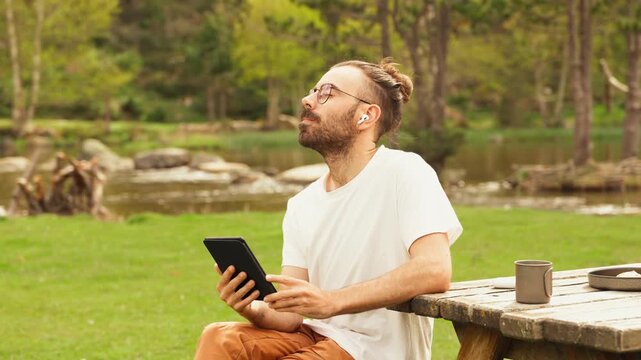 Man sits at a wooden picnic table in a green park. He uses a tablet computer, focused and relaxed. Coffee cup and frisbee rest on the table. Trees, grass, and a pond surround the scene