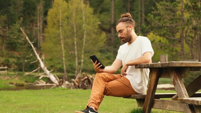 Man with a bun sits at a picnic table. He holds a tablet, calm and focused. Forest greenery surrounds him. Three keyframes capture this quiet moment