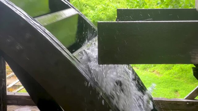 Closeup of water pouring spilling from the end of a wooden sluice turning large wooden water wheel at historic colonial American iron forge early innovation hydro power renewable resource clean energy