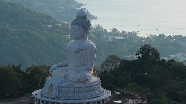 Big Buddha overlooks Phuket coastline from forested hill at golden hour, drone telephoto parallax