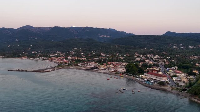 Aerial view of Roda coastal resort town in Corfu Greece at twilight with full moon over scenic mountains and Ionian sea.
