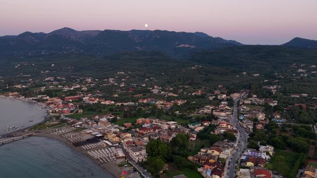 Aerial view of Roda village in North Corfu Greece at dusk with full moon rising over mountains, beautiful coastal town landscape beach sea houses roads and twilight sky