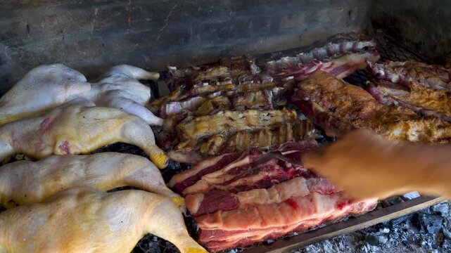 Chicken halves and thick beef ribs cook over hot coals on an Argentine asado parrilla as an asador turns the meat by hand, useful for food culture, grilling, or barbecue content.