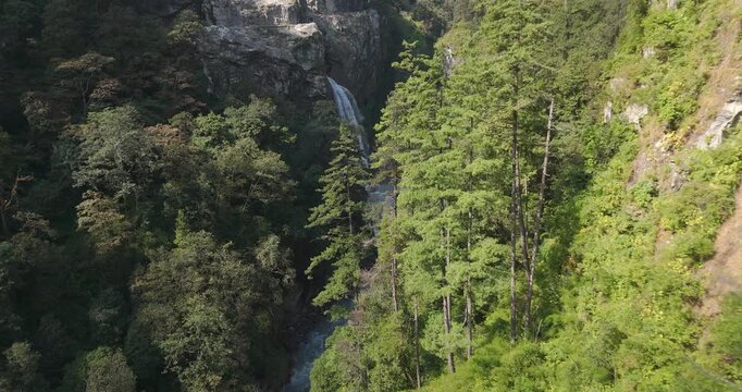 Annapurna Circuit Nepal, aerial gliding shot of Danaque waterfall cascading down a sheer cliff into the rushing turquoise Marsyangdi River gorge surrounded by dense forest in Manang.
