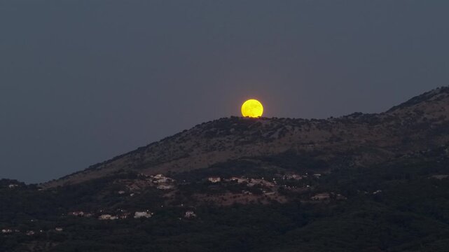 A stunning video of a glowing yellow full moon fully risen over the mountainous hills in Corfu, Greece. The deep twilight sky provides a dramatic backdrop, while the warm, scattered lights of a small